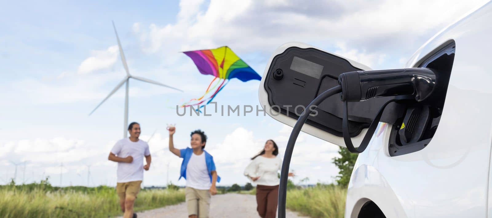 Concept of progressive happy family holding windmill toy and relax at wind farm with electric vehicle. Electric vehicle driven by clean renewable energy from wind turbine generator to charger station.