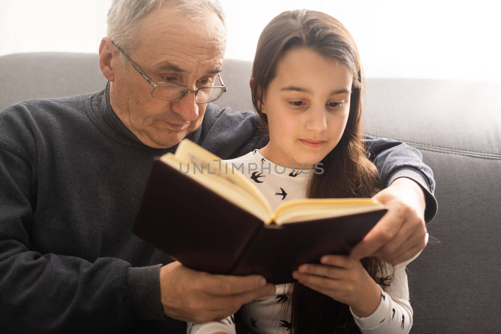 Photo of aged old grandpa little pretty granddaughter sit sofa hugging stay house quarantine safety read interesting book fairy tale modern interior living room indoors