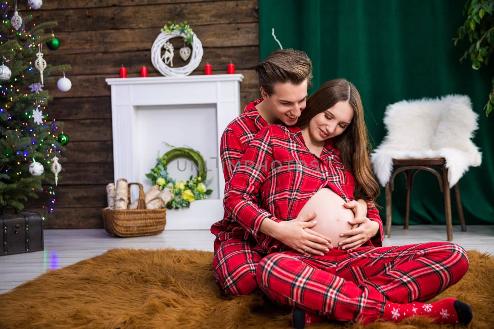 The parents-to-be hug on the carpet. The man sits behind the woman. The woman lies down and leans her back against the man. The woman is advanced in pregnancy and exposes her belly.