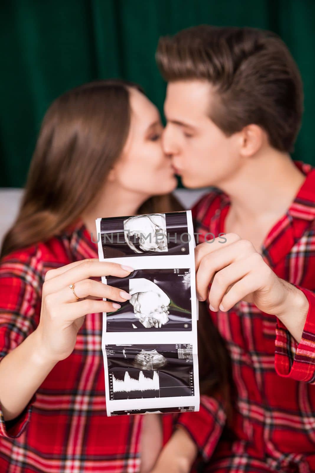 A close-up of an usg photo of a baby in the foreground. The photo is held by the parents-to-be visible in the background. Blurred second plan. In the background a woman and a man kissing