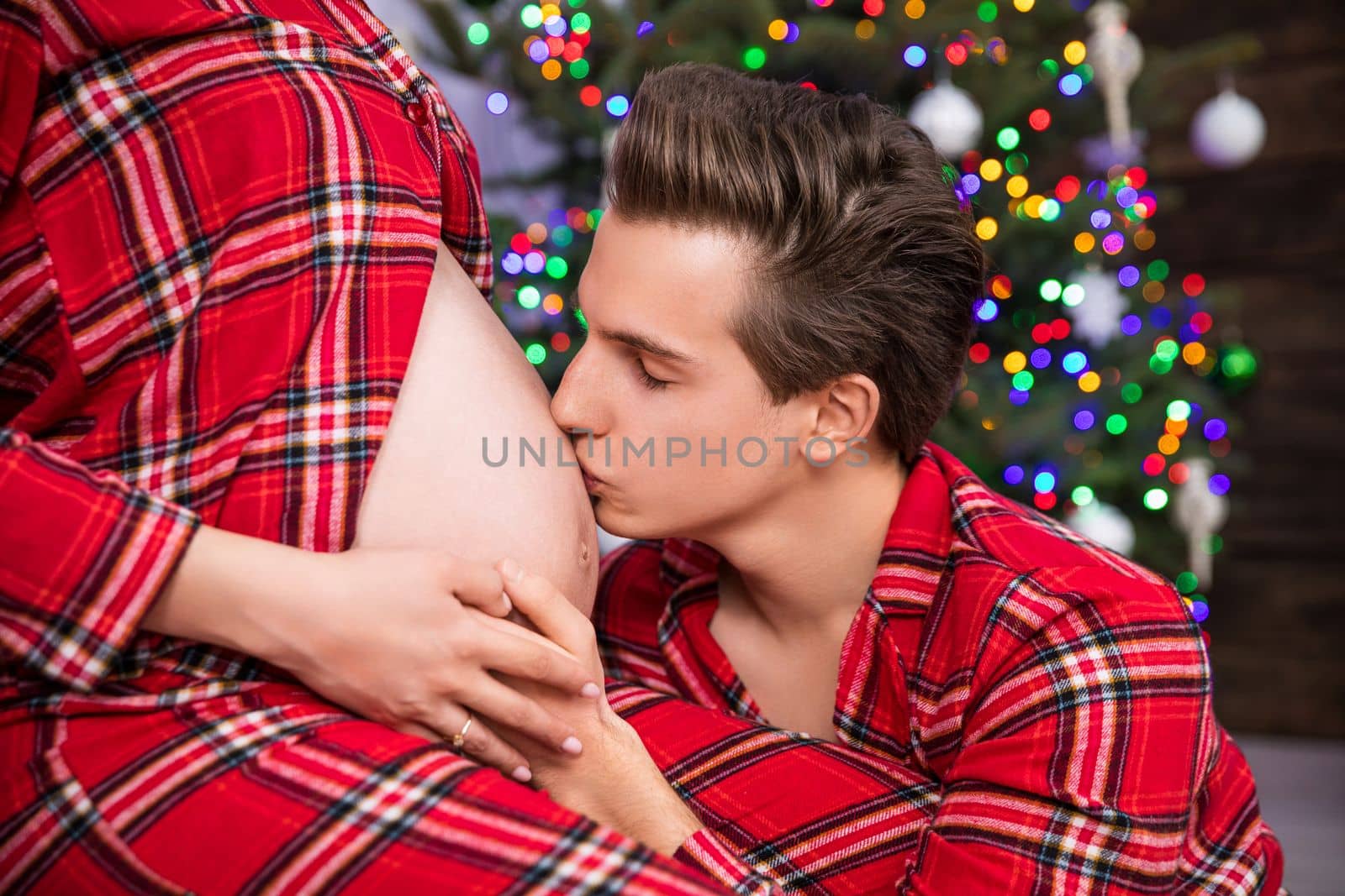 Against a background of Christmas decorations and a Christmas tree, a husband kneels in front of his wife. Close-up on the profile of the pregnant belly that the kneeling man is kissing. The partners hold hands on the pregnant belly.