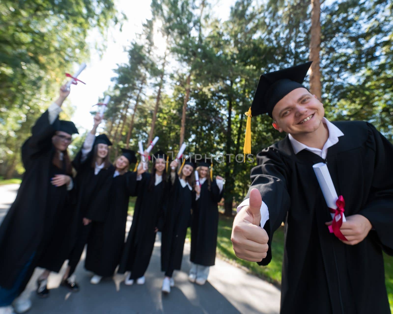 Happy young caucasian male graduate showing thumbs up. A group of graduate students outdoors. by mrwed54