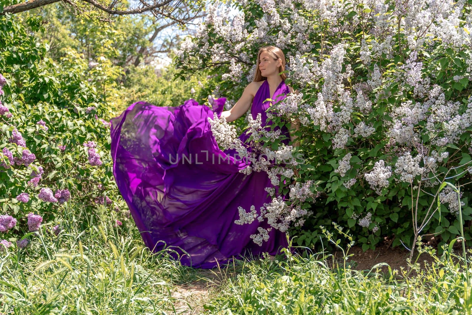 Fashion Model in Lilac Flowers, Young Woman in Beautiful Long Dress Waving on Wind, Outdoor Beauty Portrait in Blooming Garden by Matiunina