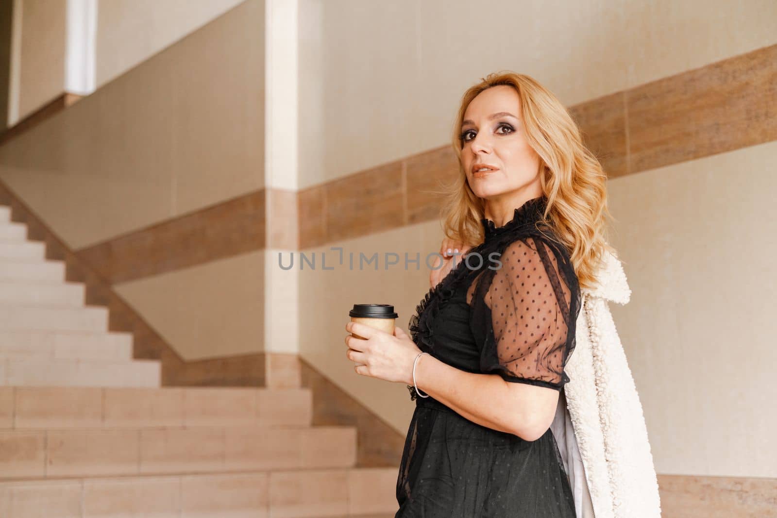 Portrait of a beautiful smiling blonde. She's walking up the stairs, coffee in hand. The Concept Of Urban Life. by Matiunina