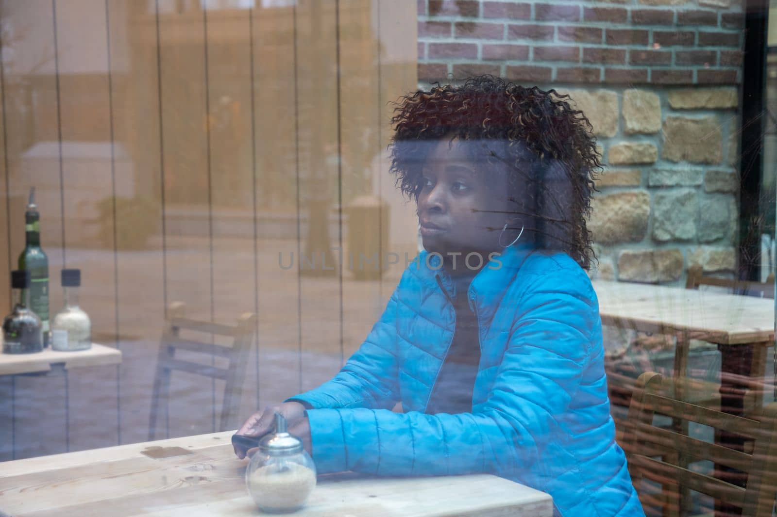 An African American woman in a blue jacket sits behind a window glass in a cafe waiting for a waiter, selective soft focus of the photo due to dirty glass. High quality photo