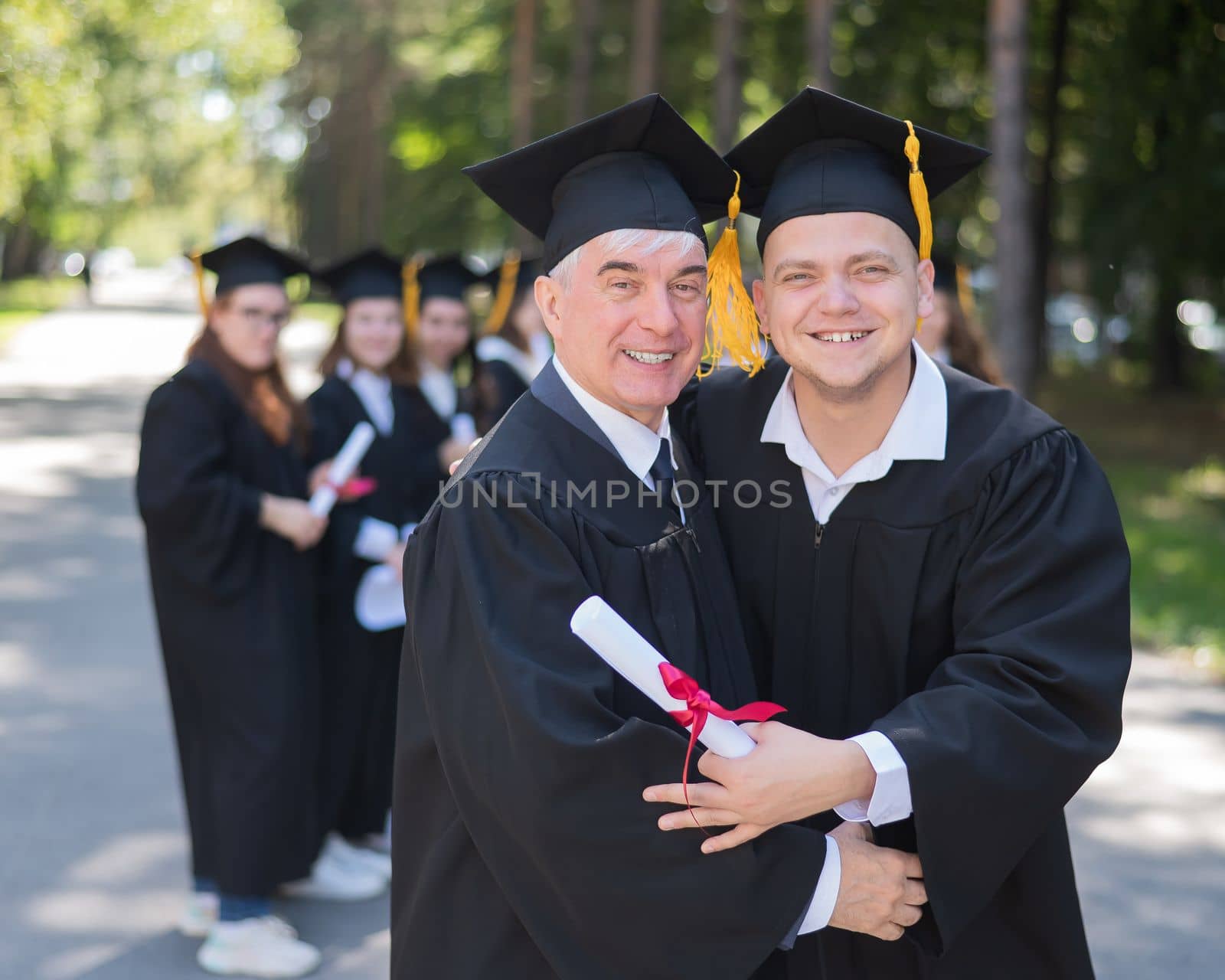 A group of graduates in robes outdoors. An elderly man and a young guy congratulate each other on receiving a diploma. by mrwed54