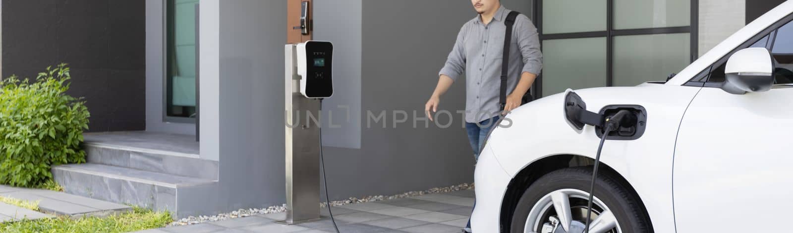 Progressive asian man and electric car with home charging station. Concept of the use of electric vehicles in a progressive lifestyle contributes to a clean and healthy environment.