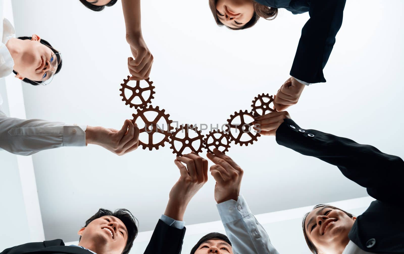 Hand holding wooden gear by businesspeople wearing suit for harmony synergy in office workplace. Bottom view office worker hand make chain of gear into collective unity form.