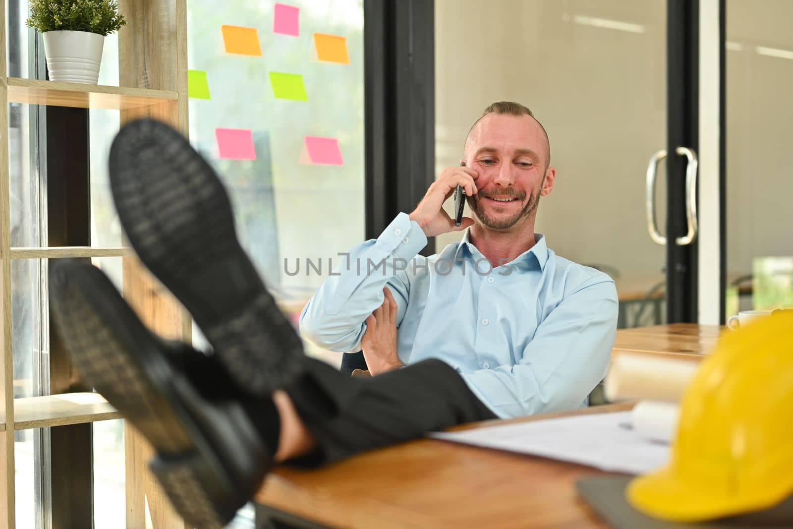 Pleasant caucasian engineer in shirt putting legs on table and talking on mobile phone in his personal office.