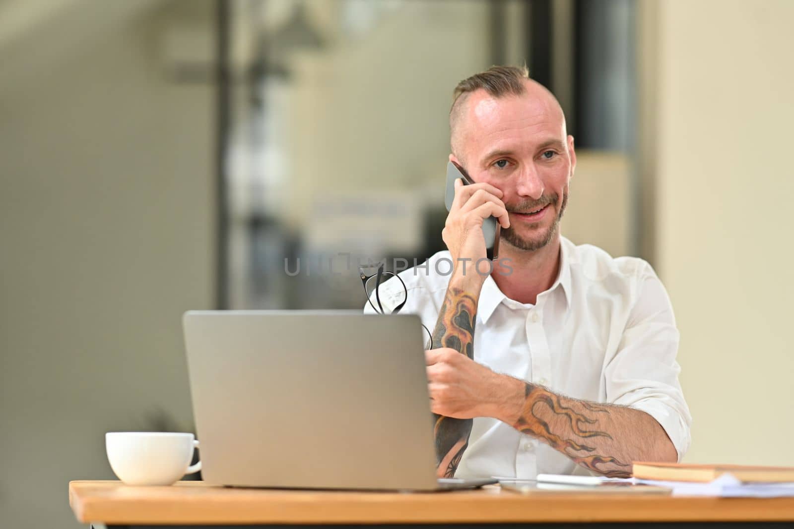 Confident caucasian male manager in a white shirt with tattooed sitting in front of laptop at office desk and talking on mobile phone.