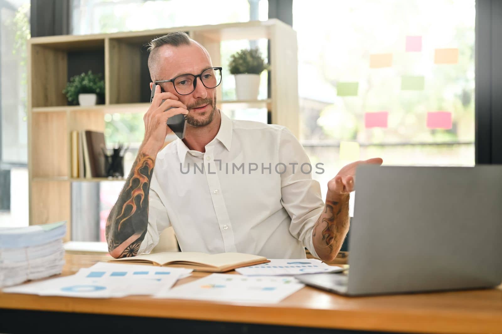 Portrait of hipster businessman having pleased phone conversation and using laptop computer in bright office.