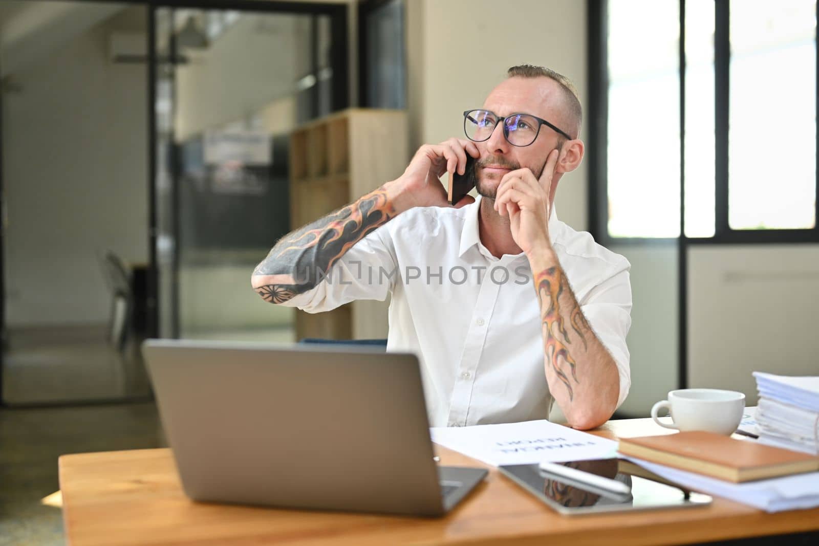Pleasant tattooed businessman in white shirt having phone conversation and looking through office window.