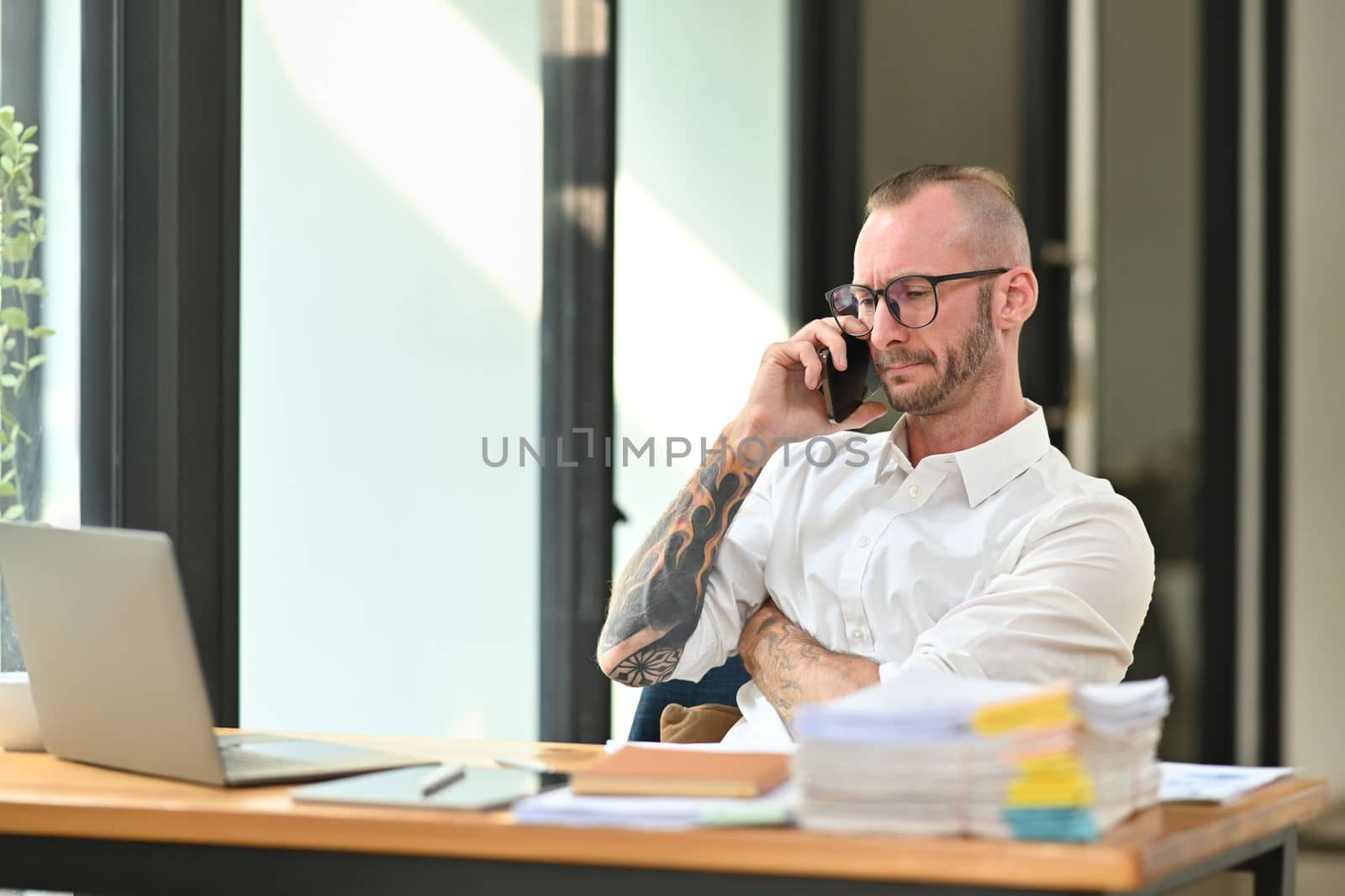 Confident handsome businessman in a white shirt with tattooed talking mobile phone and using laptop at his workplace by prathanchorruangsak