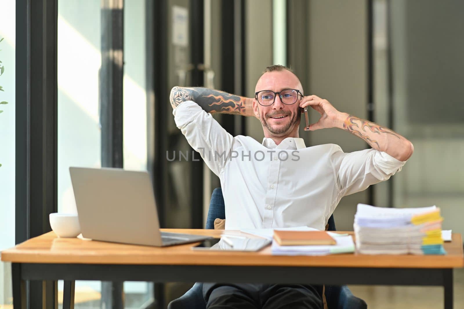 Joyful tattooed businessman having phone conversation while sitting in bright office by prathanchorruangsak