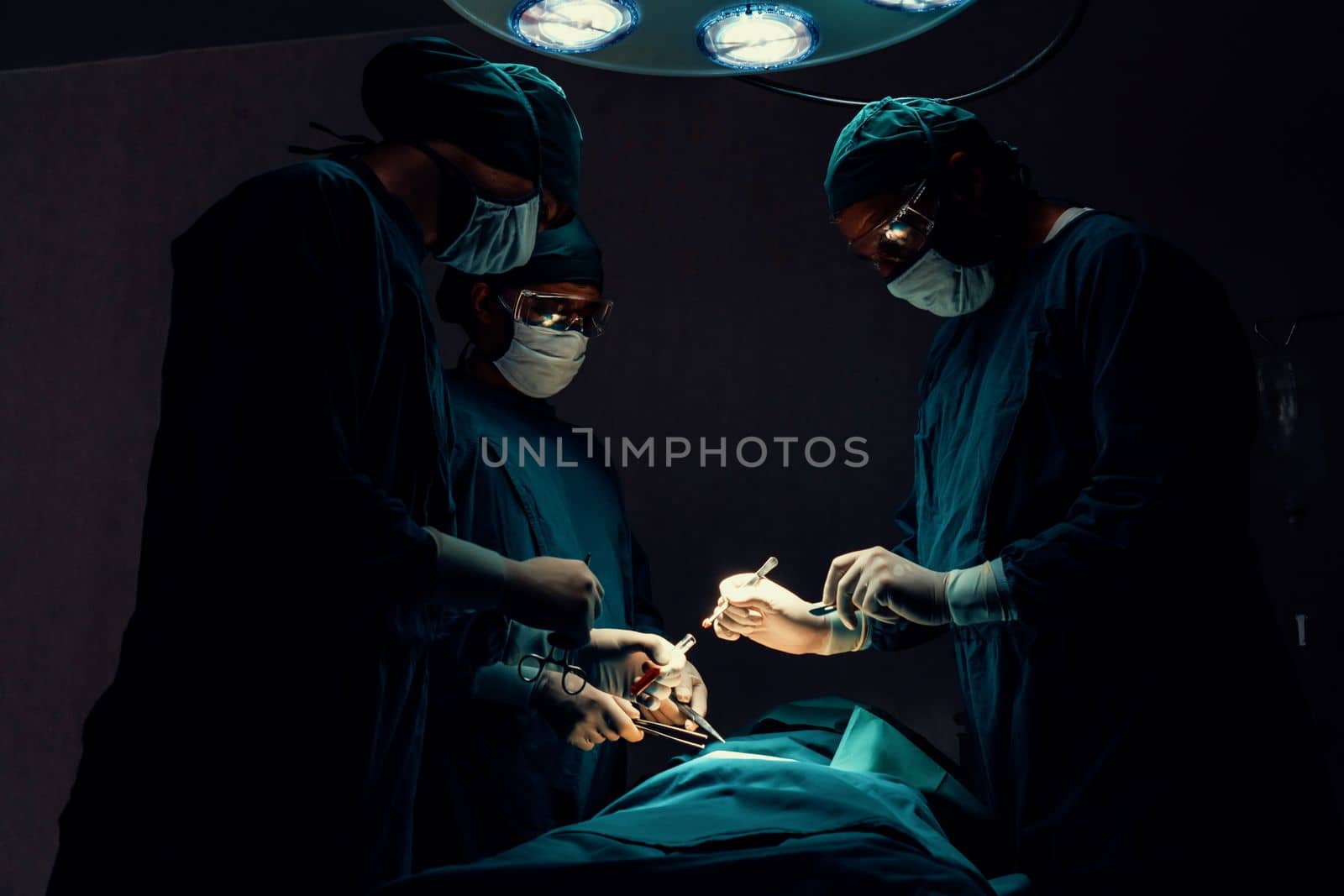 Surgical team performing surgery to patient in sterile operating room. In a surgery room lit by a lamp, a professional and confident surgical team provides medical care to an unconscious patient.