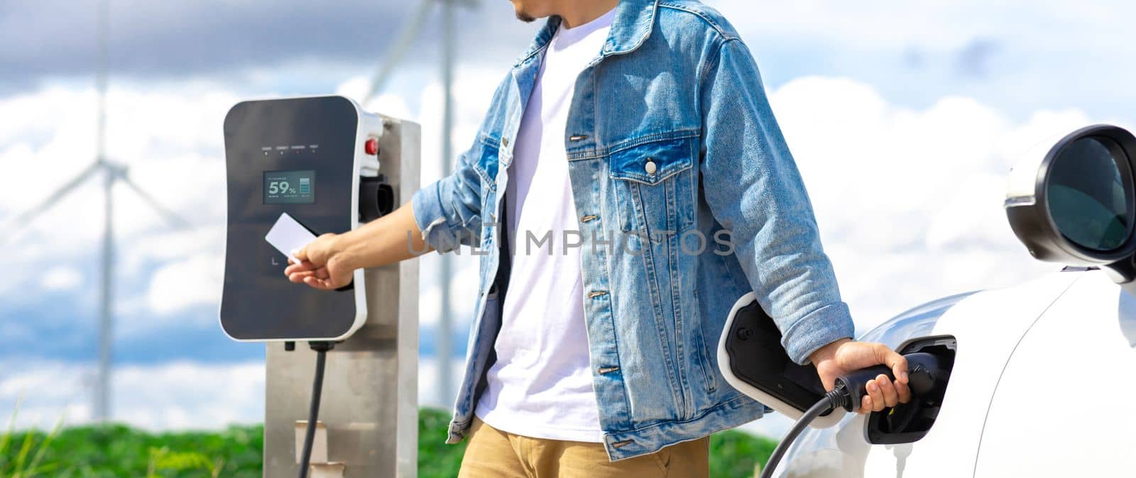 Progressive man with his electric car, EV car recharging energy from charging station on green field with wind turbine as concept of future sustainable energy. Electric vehicle with energy generator.