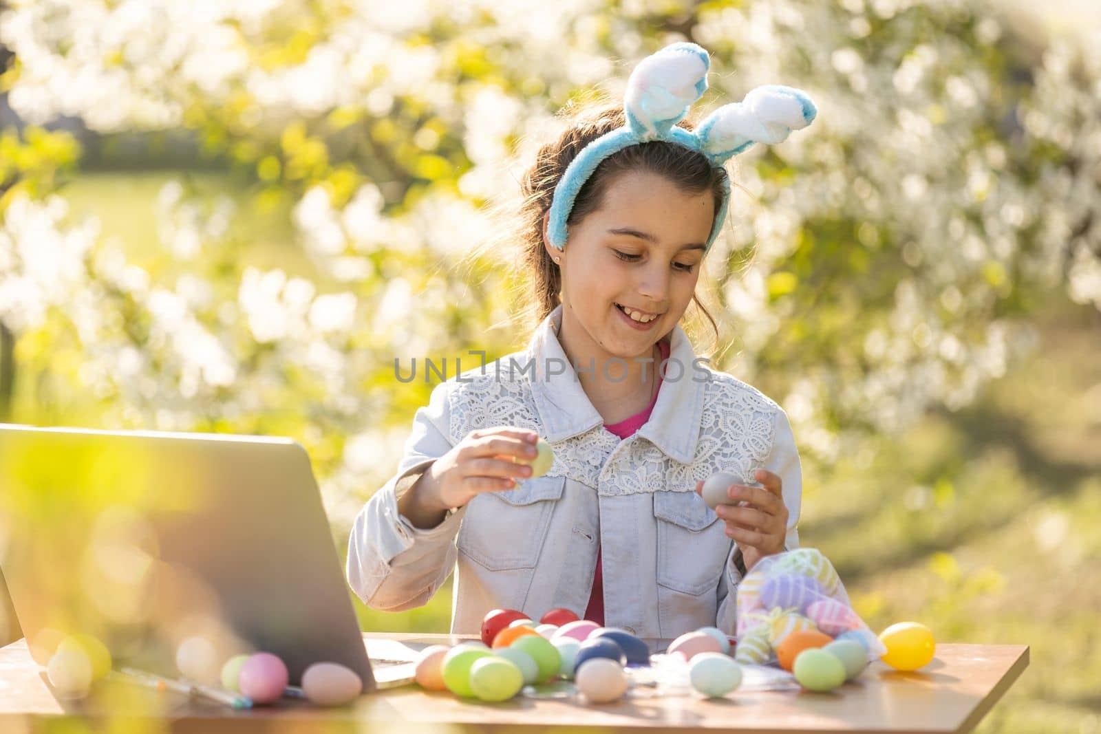 happy teen girl wear bunny ears paints Easter eggs with a laptop in the garden by Andelov13