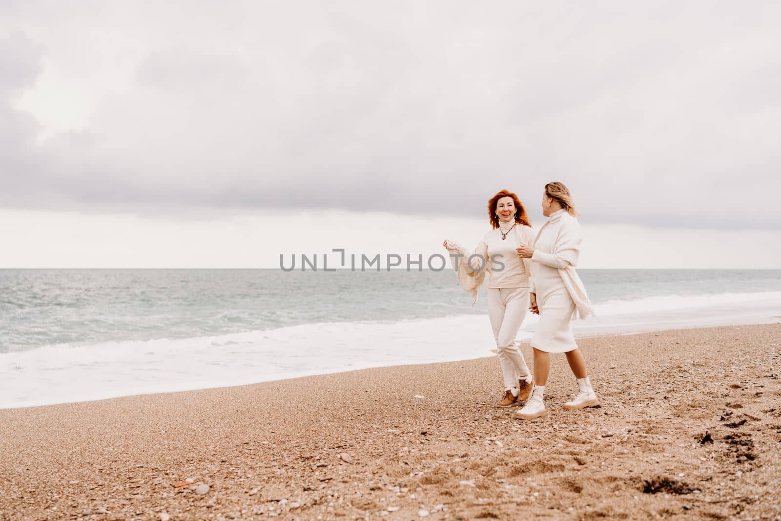 Women sea walk friendship spring. Two girlfriends, redhead and blonde, middle-aged walk along the sandy beach of the sea, dressed in white clothes. Against the backdrop of a cloudy sky and the winter sea. Weekend concept. by Matiunina