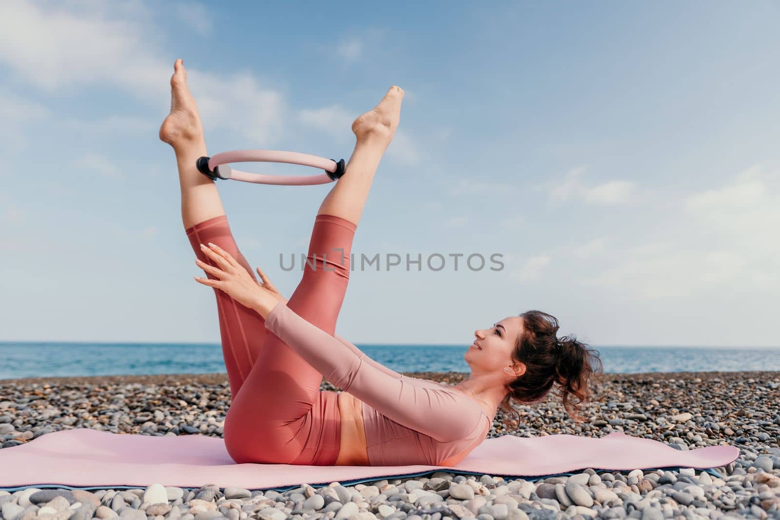 Middle aged well looking woman with black hair doing Pilates with the ring on the yoga mat near the sea on the pebble beach. Female fitness yoga concept. Healthy lifestyle, harmony and meditation.