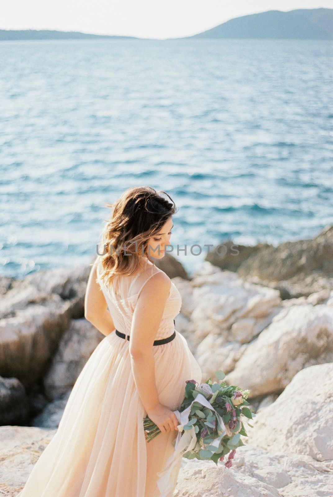 Bride with a bouquet stands sideways with her head bowed on a rocky seashore. High quality photo