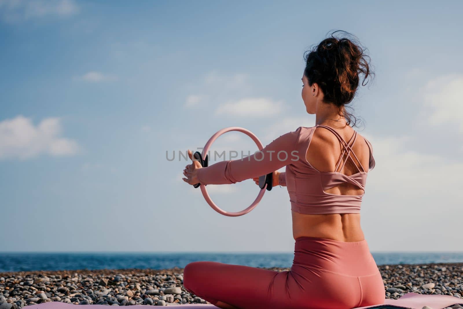Middle aged well looking woman with black hair doing Pilates with the ring on the yoga mat near the sea on the pebble beach. Female fitness yoga concept. Healthy lifestyle, harmony and meditation.