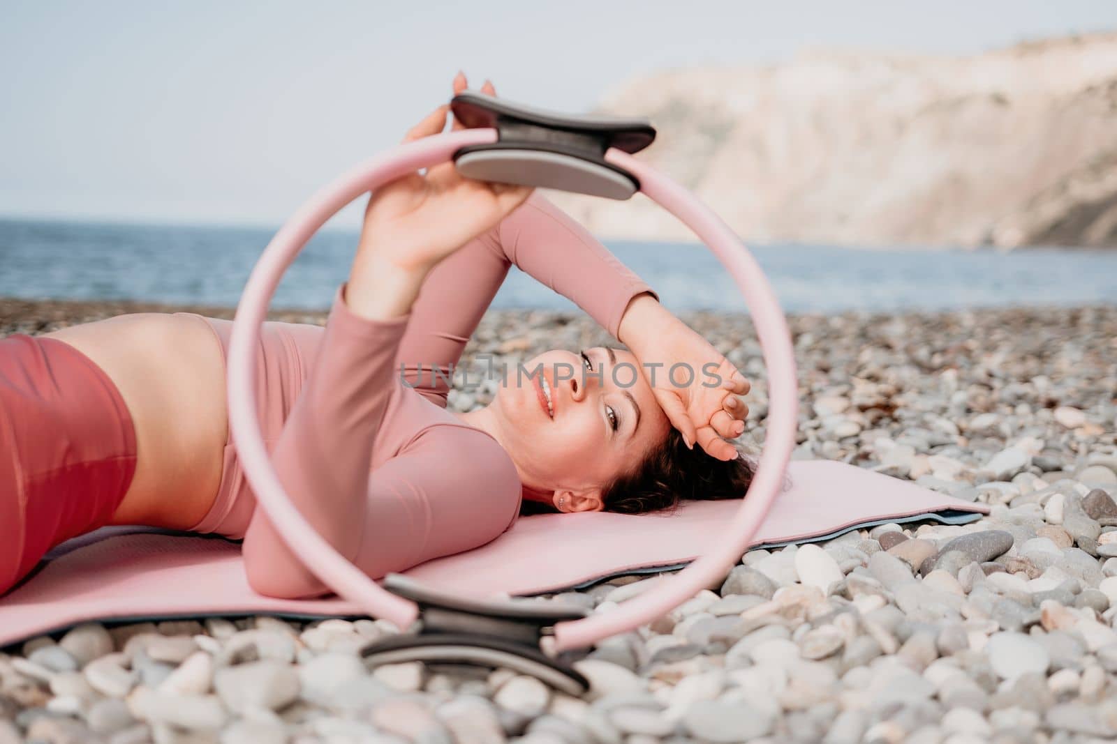 Middle aged well looking woman with black hair doing Pilates with the ring on the yoga mat near the sea on the pebble beach. Female fitness yoga concept. Healthy lifestyle, harmony and meditation.