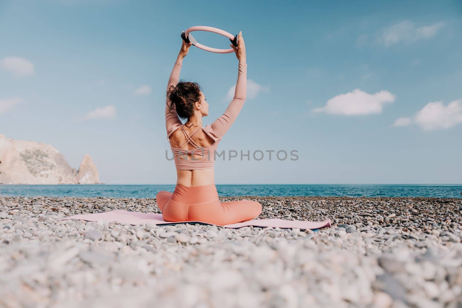 Middle aged well looking woman with black hair doing Pilates with the ring on the yoga mat near the sea on the pebble beach. Female fitness yoga concept. Healthy lifestyle, harmony and meditation.