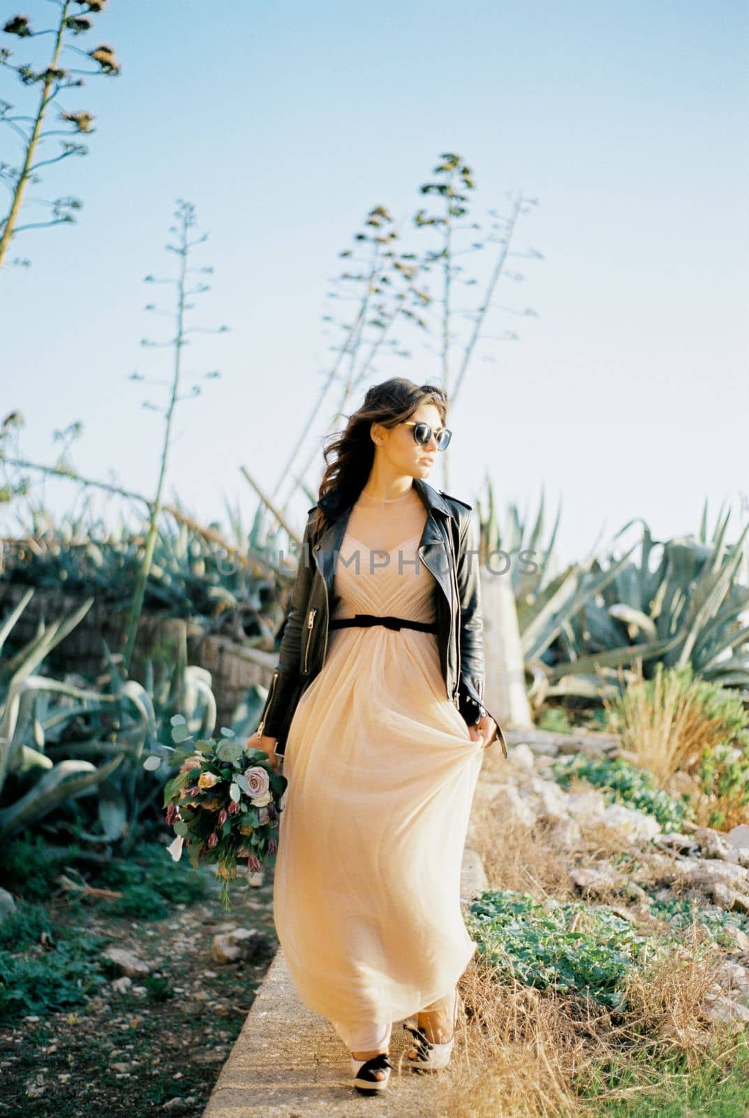 Bride in sunglasses walks along a stone curb with her head turned to the side. High quality photo