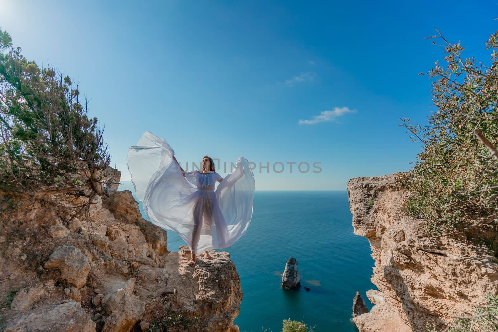 A beautiful young woman in a white light dress with long legs stands on the edge of a cliff above the sea waving a white long dress, against the background of the blue sky and the sea