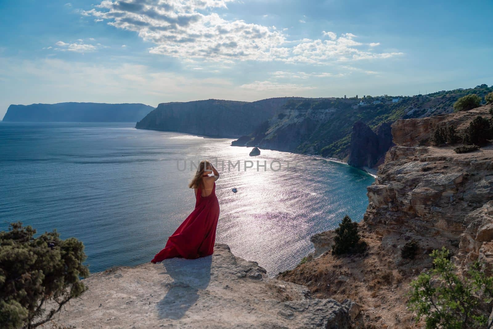 A girl with flowing hair in a long red dress stands on a rock above the sea. The stone can be seen in the sea. Sunny path to the sea from the sun