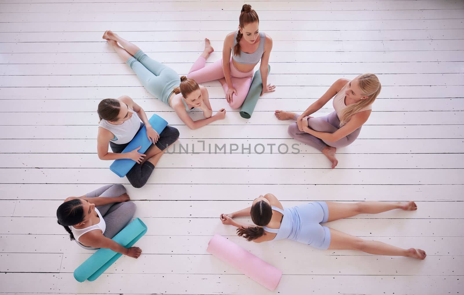Group of women relaxing in yoga class from above. Friends talking, being social in pilates class. Women bonding in yoga class. Young women sitting on the floor in yoga class from above by YuriArcurs