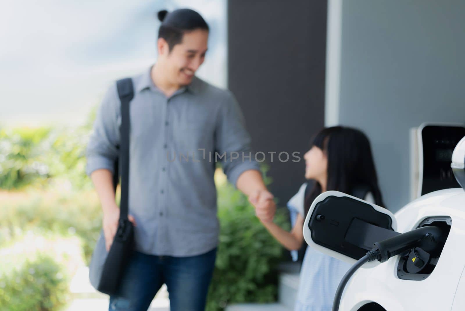 Focus electric car recharging at home charging station with blurred father and daughter walking in background. Progressive green and clean energy vehicle for healthy environment lifestyle concept.