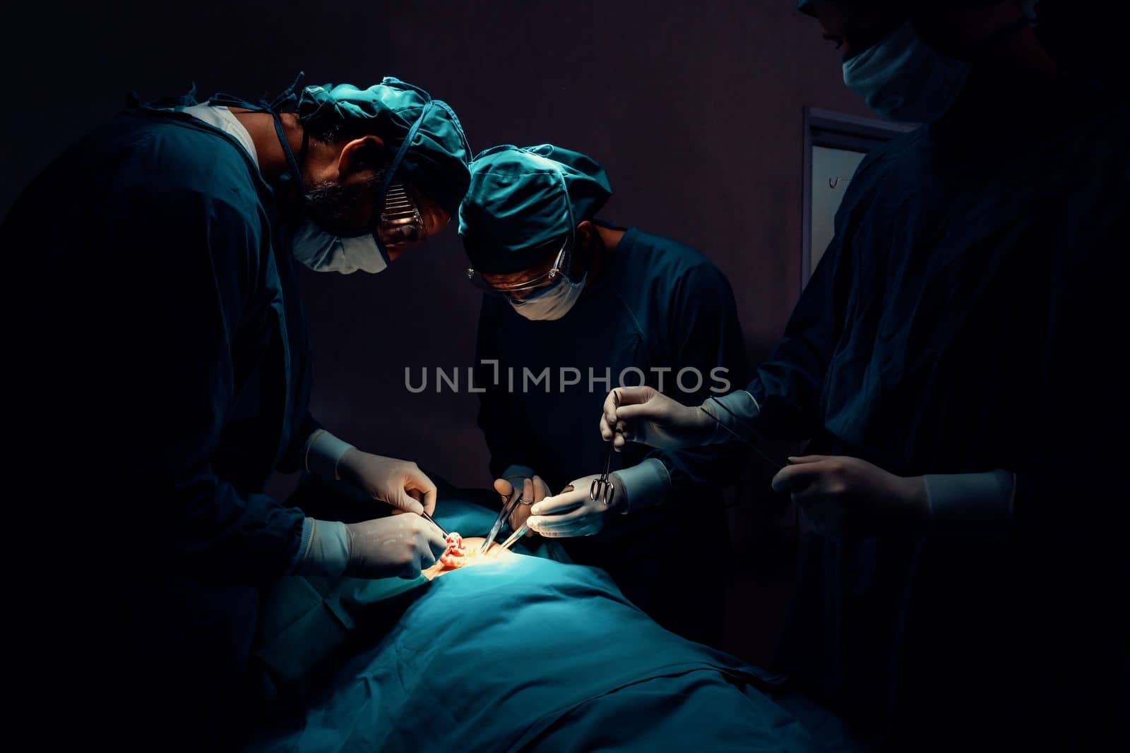 Surgical team performing surgery to patient in sterile operating room. In a surgery room lit by a lamp, a professional and confident surgical team provides medical care to an unconscious patient.