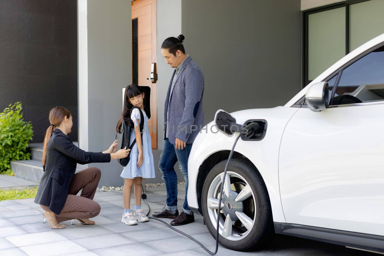 Progressive young parents and daughter with electric vehicle and home charging station. Green and clean energy from electric vehicles for healthy environment. Eco power from renewable source at home.