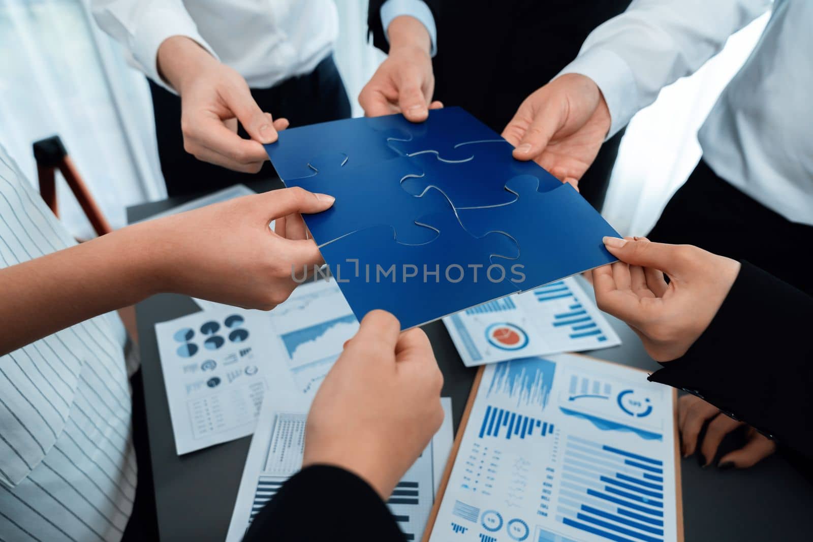 Closeup top view business team of office worker putting jigsaw puzzle together over table filled with financial report paper in workplace with manager to promote harmony concept in meeting room.