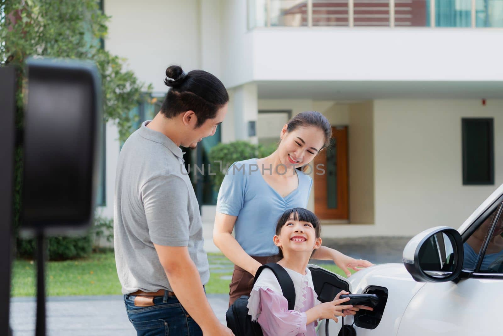 Progressive young parents and daughter with electric vehicle and home charging station. Green and clean energy from electric vehicles for healthy environment. Eco power from renewable source at home.