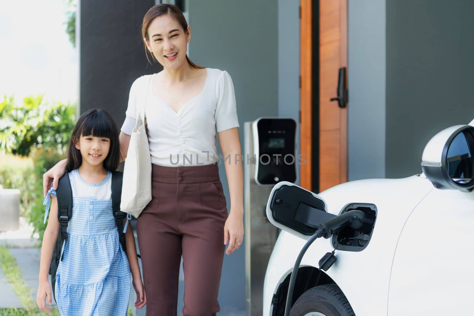 Progressive lifestyle of mother and daughter who have just returned from school in an electric vehicle that is being charged at home. Electric vehicle powered by sustainable clean energy.