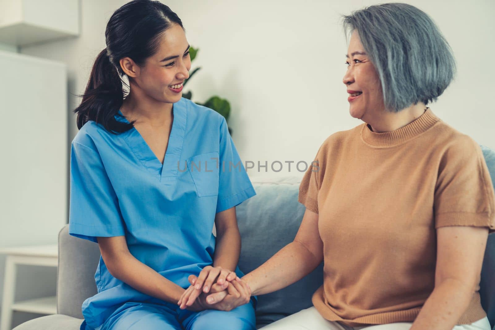 A caregiver rest her hands on the shoulders of a contented senior patient while she sitting on the sofa at home.