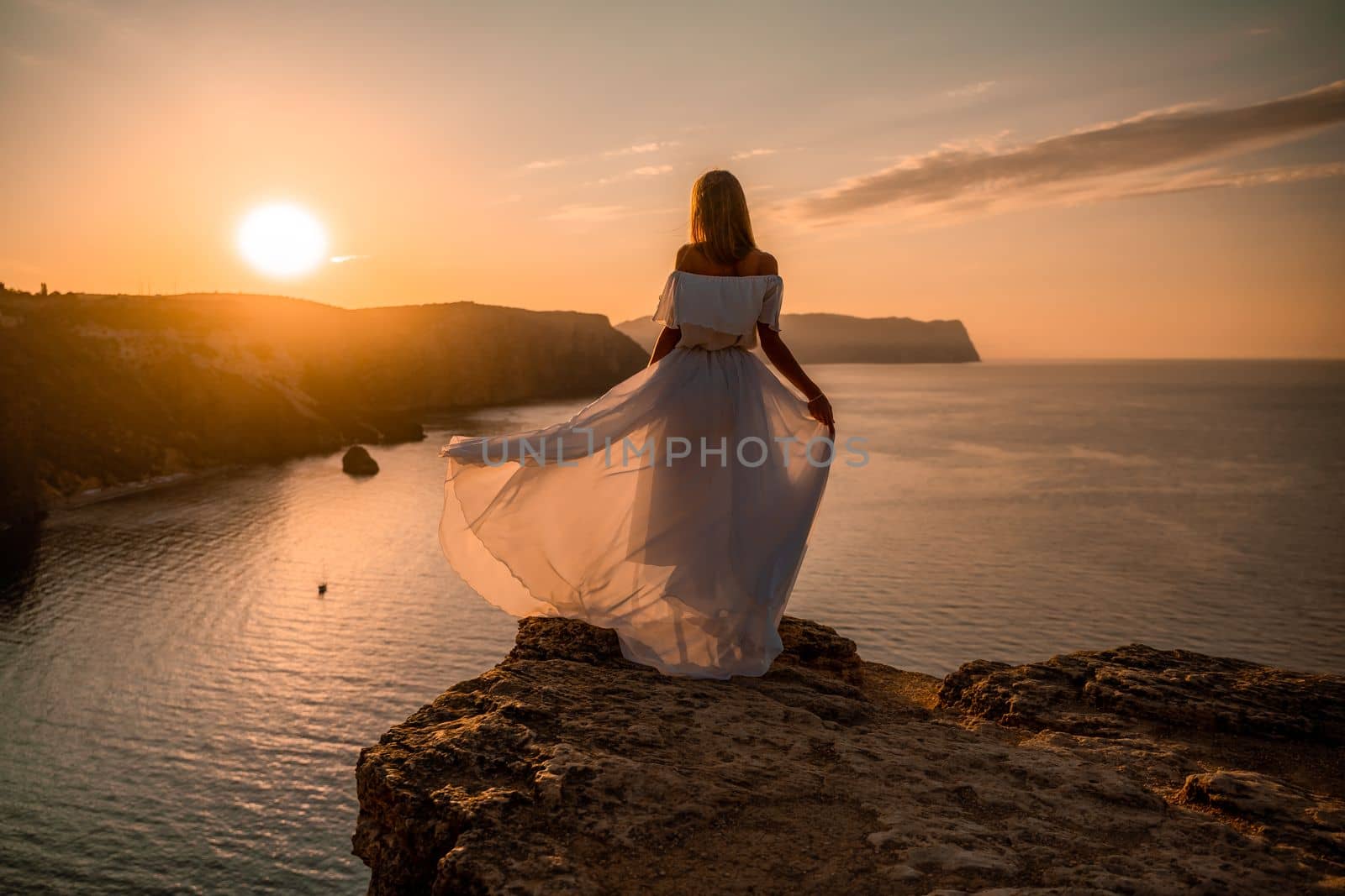Sunset woman dress. A happy woman in a white light dress stands on the edge of a cliff above the sea, waving a white long dress, against the background of the sunrise of the sun and the sea