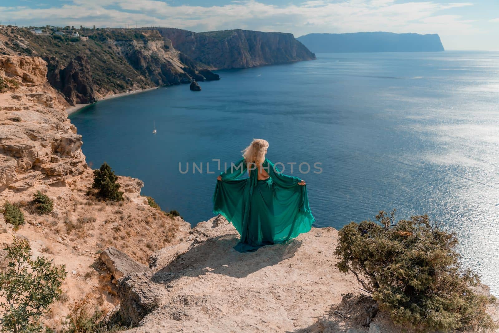 A beautiful young woman in a mint light dress with long legs stands on the edge of a cliff above the sea waving long dress, against the background of the blue sky and the sea