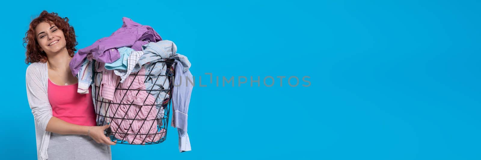 A girl with a smile on her face carries a basket full of washed and dried men's shirts. Joy and contentment. Dark curly hair. . Panoramic view of girl with empty blue background.