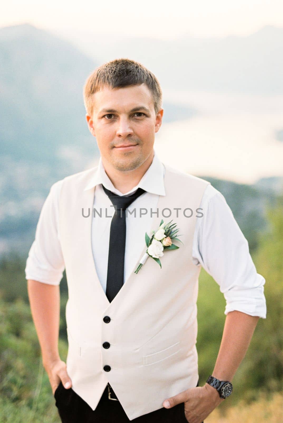 Groom in a vest with a boutonniere stands on a mountain with his hands in his pockets. High quality photo