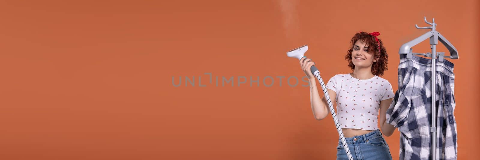 Hot steam used for ironing clothes. Steam device. An ironing steamer used by a young woman. Panoramic view of girl with empty orange background.