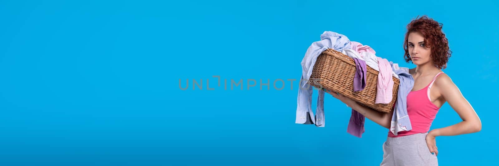 A woman with an angry face carries a wicker basket with shirts for washing. Daily household chores. Young attractive girl with curly hair.
