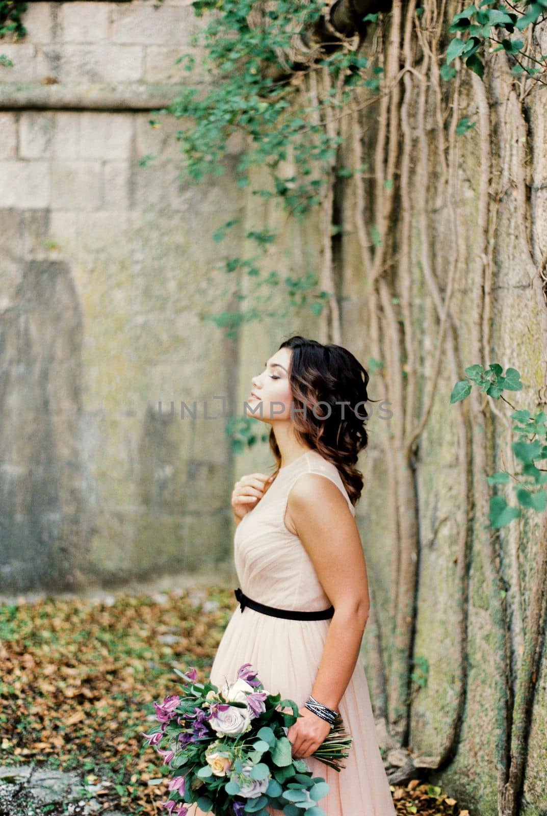 Bride with a bouquet stands near a stone wall entwined with ivy. High quality photo