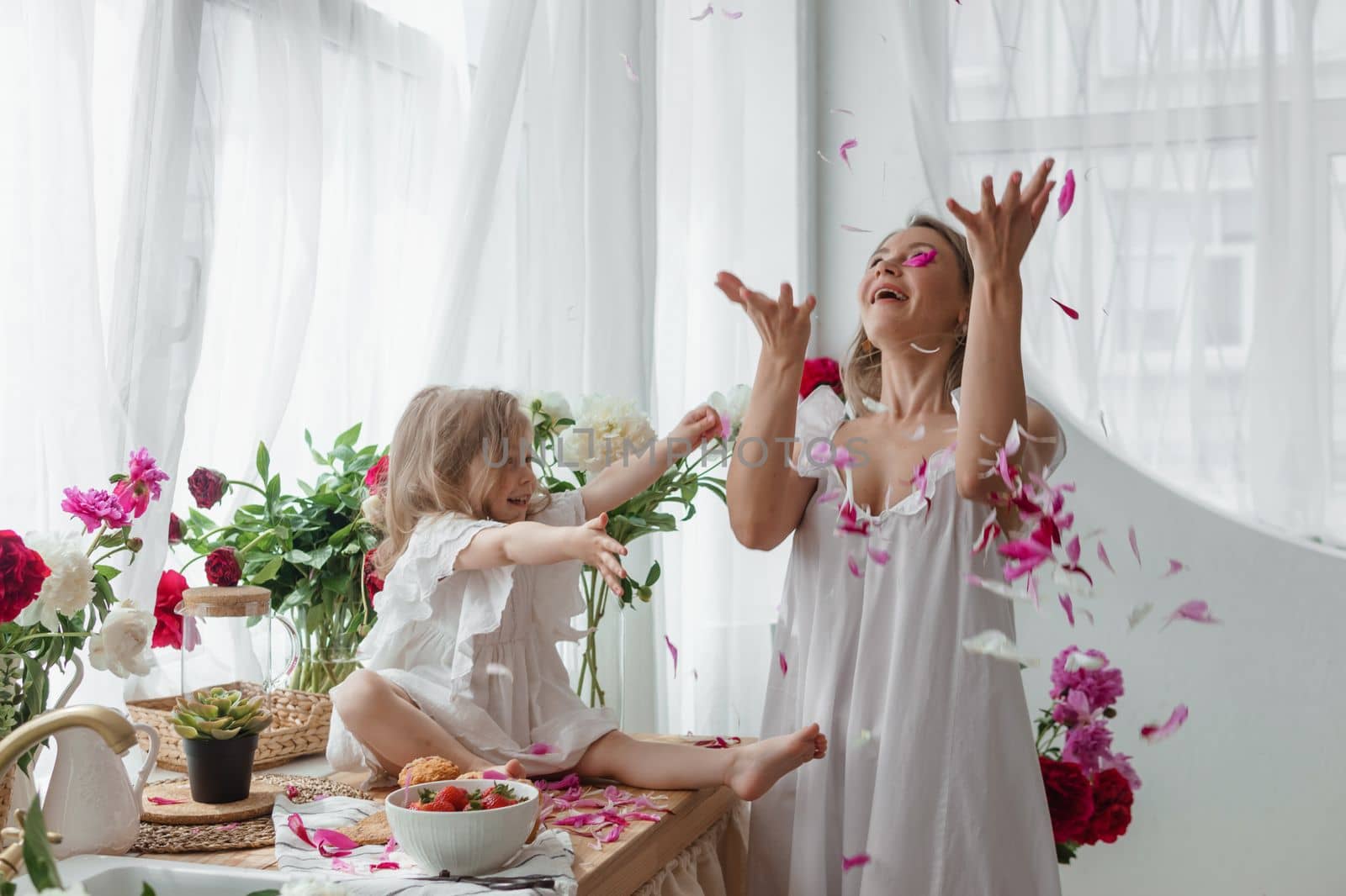 A little blonde girl with her mom on a kitchen countertop decorated with peonies. The concept of the relationship between mother and daughter. Spring atmosphere.