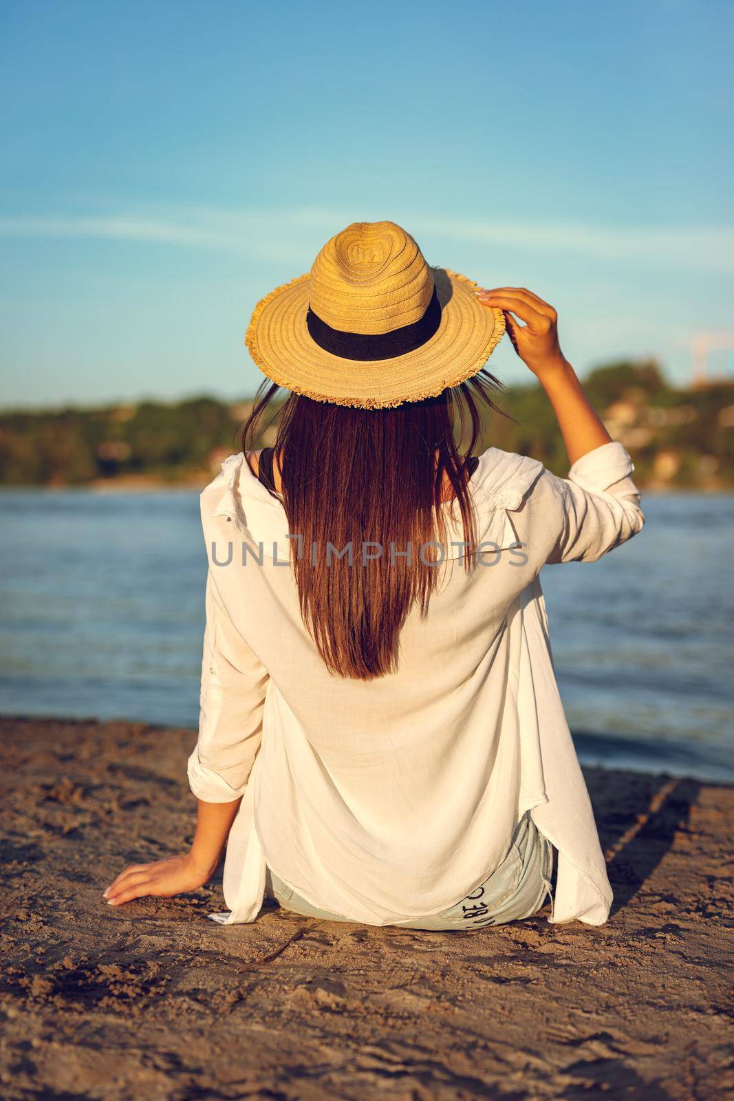 Rear view on a young woman in straw hat relaxing at sunset time on the river bank.