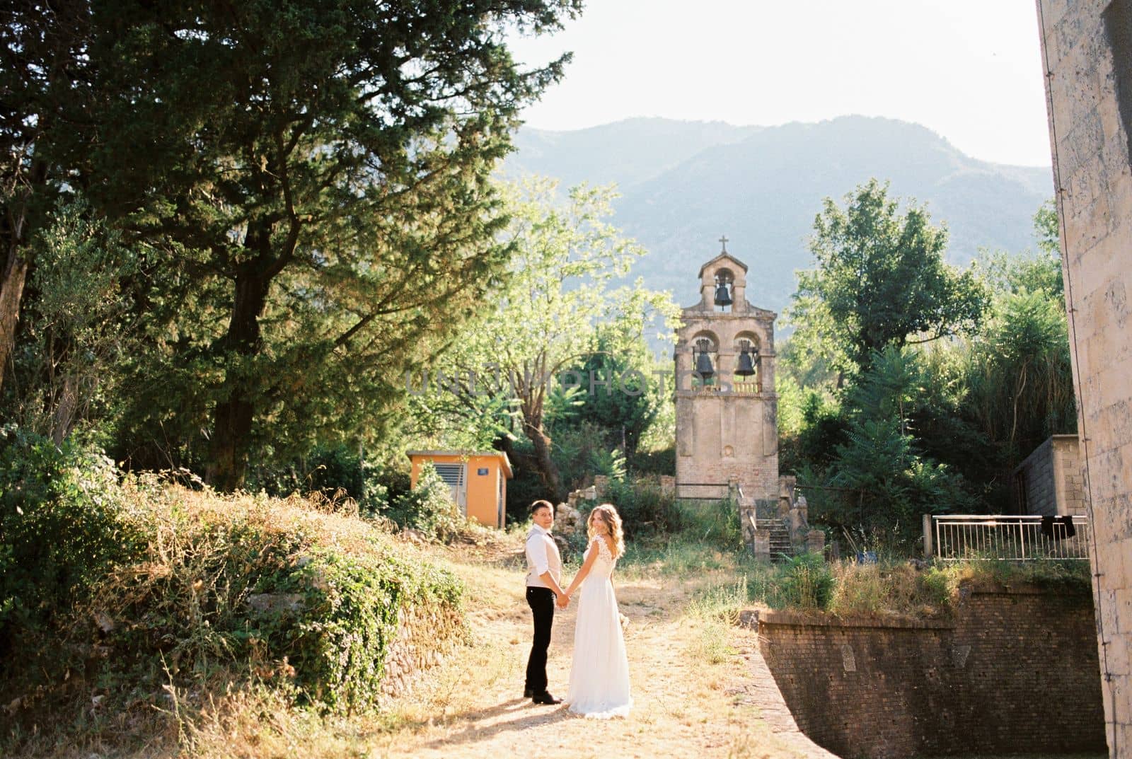 Bride and groom stand holding hands on the path to the ancient chapel. High quality photo