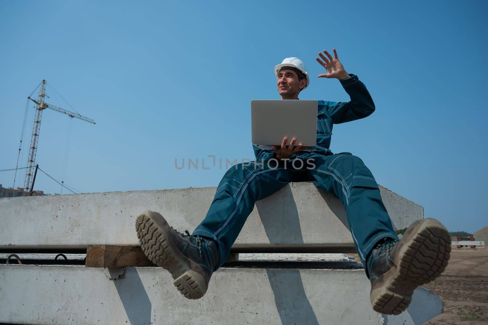 Caucasian male builder in hardhat sits on floor slabs and uses laptop at construction site