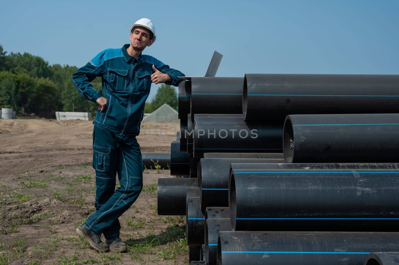 Caucasian male builder in a hard hat stands near the pipes and uses a laptop at a construction site